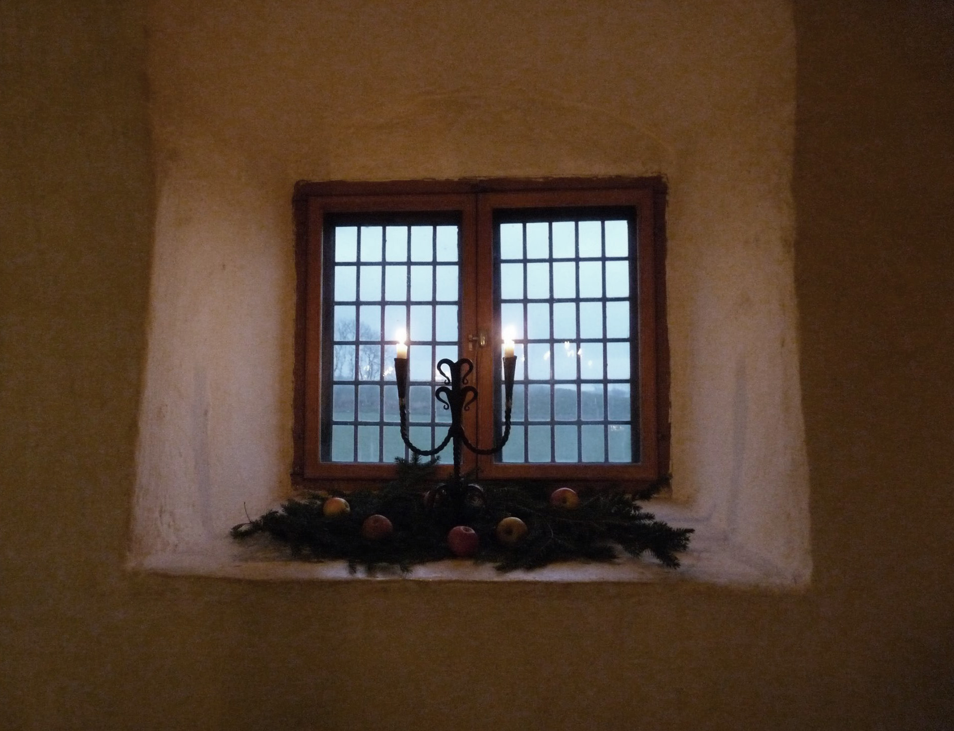 Ett djupt sittande fönster i en vitputsad vägg. I fönstret ligger granris med röda äpplen och i en ljusstake brinner ett ljus.