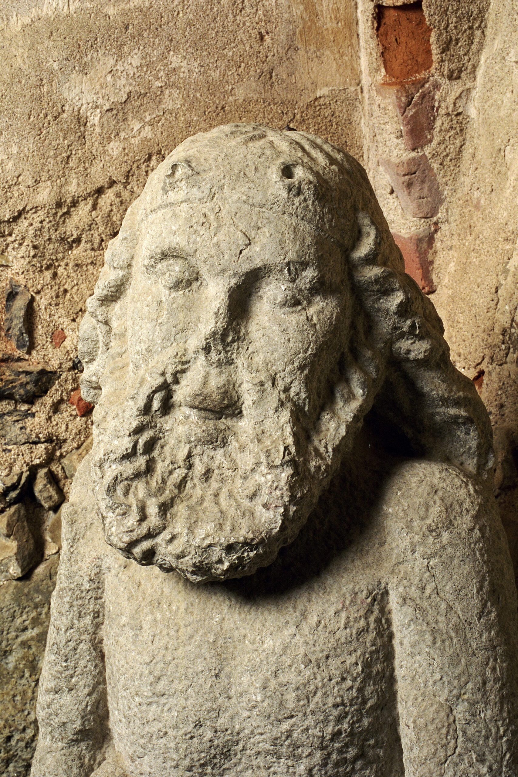 Stenstatyn Vildmannen ses från bröstet och upp. Han har axellångt skägg och hår och en lite bortslagen näsa. Direkt bakom ses borgens vägg med kalkputs och tegel.