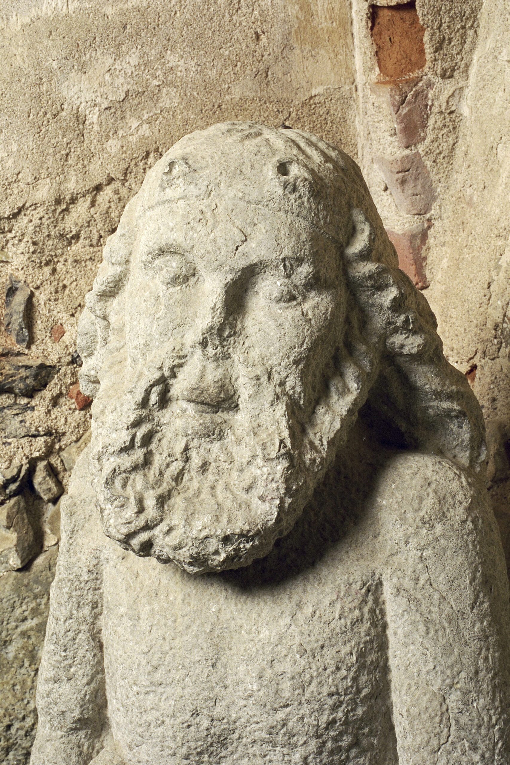 The stone statue of the Wild Man is seen from the chest up. He has shoulder-length beard and hair and a slightly knocked-out nose. Directly behind you can see the castle's wall with lime plaster and brick.