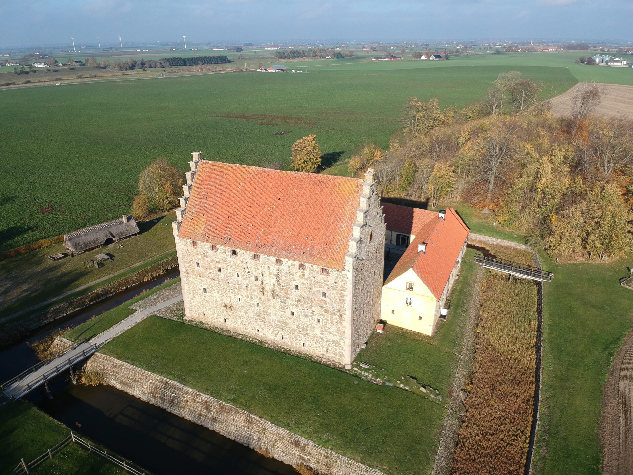 The castle seen from above from a drone. A square moat surrounds the castle yard, where two of the houses can also be seen. To the left, green fields.