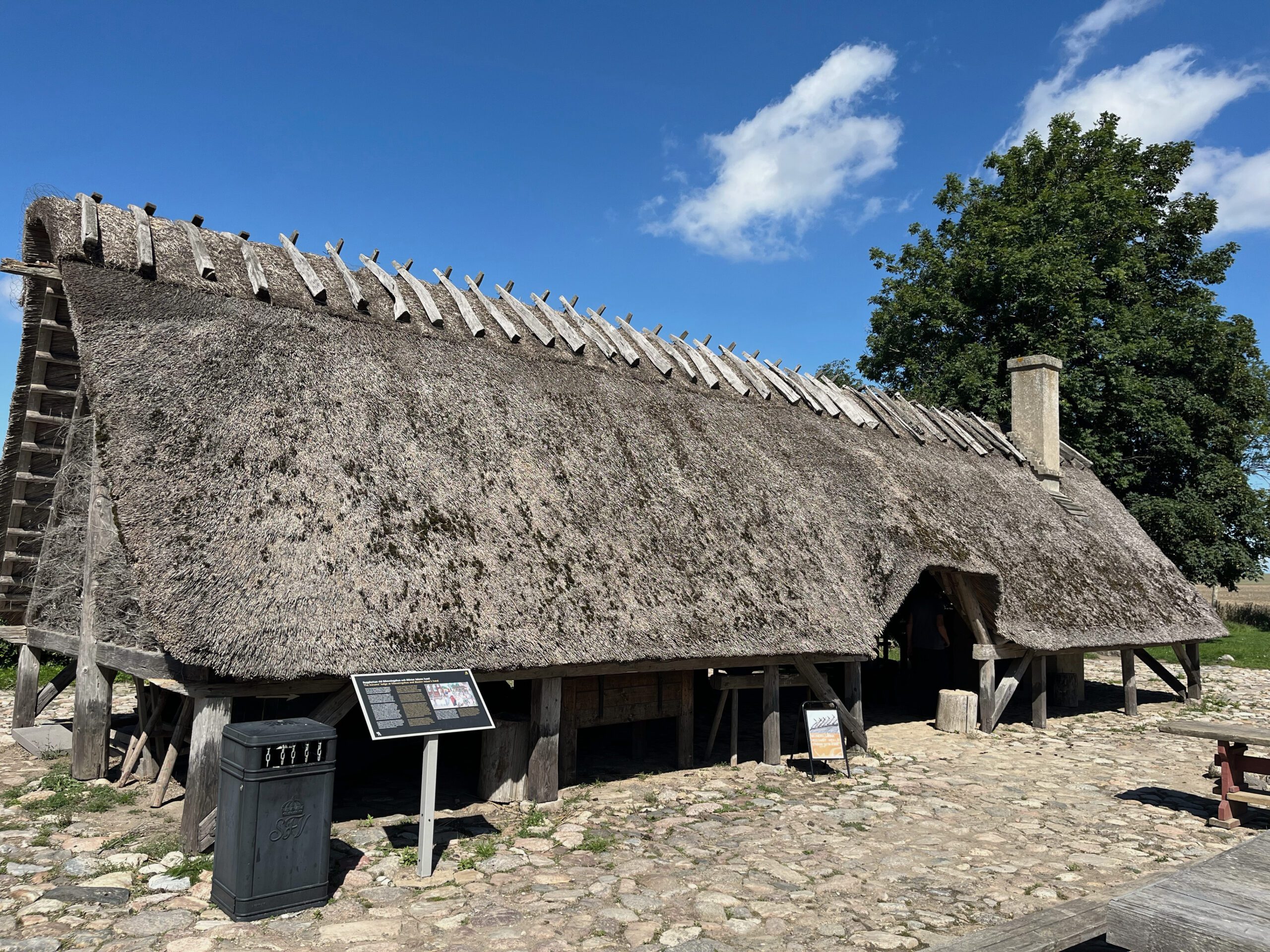 A long, low building with a thatched roof and open walls. A chimney is on the right side of the roof. The ground in front is paved with cobblestones. A tree stands diagonally behind on the right.