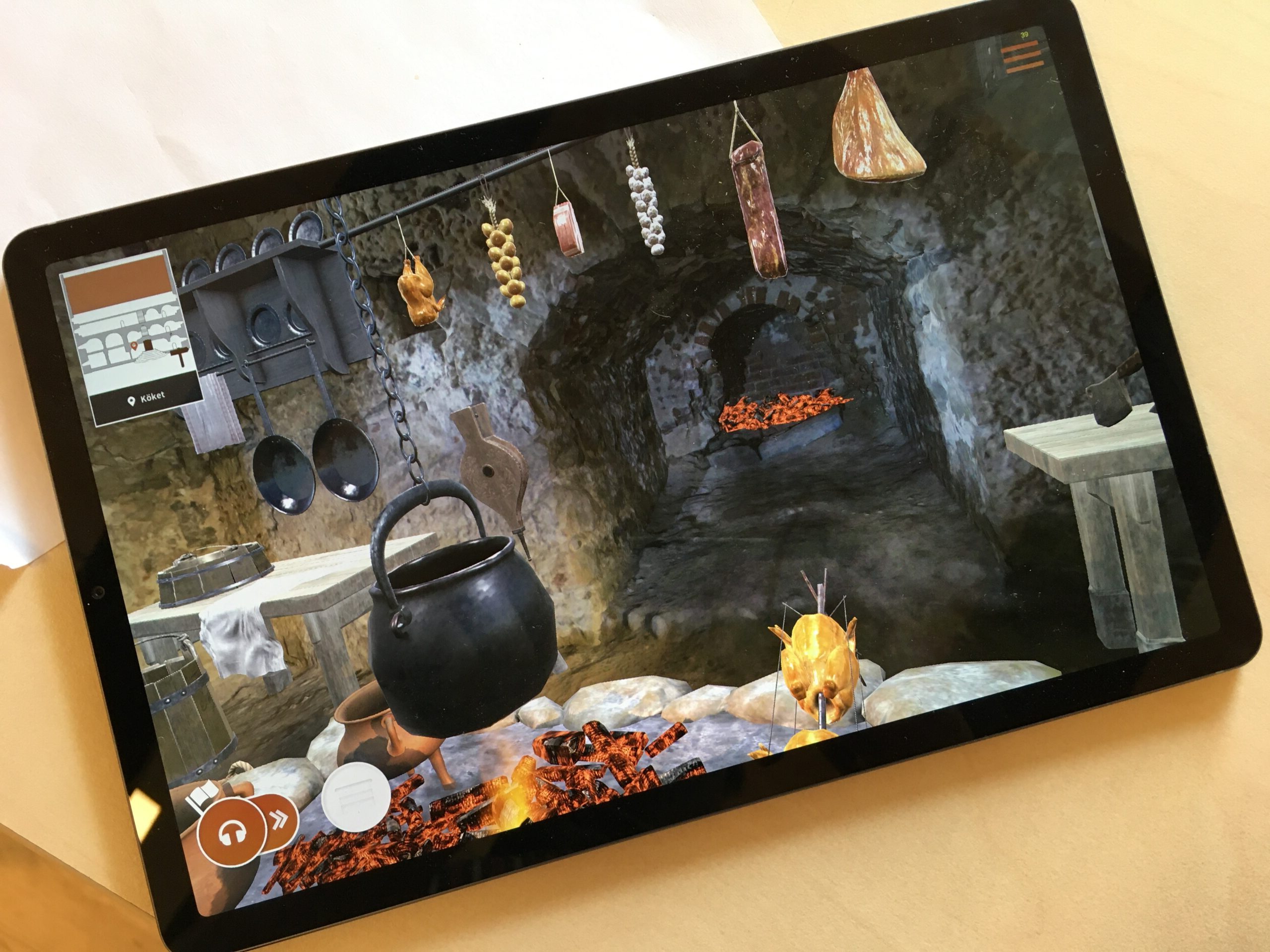 A tablet shows a 3D model of the castle's kitchen, as it might have looked. Onions, chicken and ham hang from a pole in the ceiling. A pot hangs over a glowing hearth.