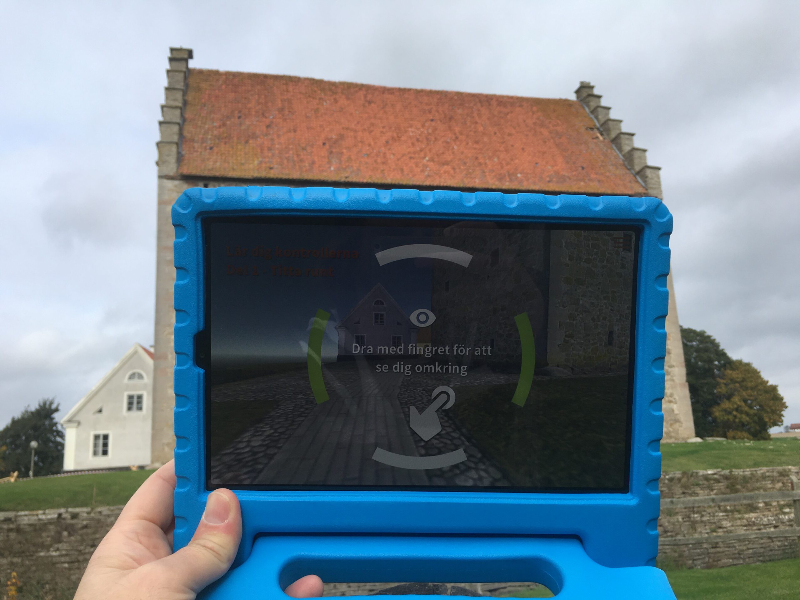 A tablet with the digital guide on the screen is held up outside in front of the castle so that the screen covers the entire building except the roof.