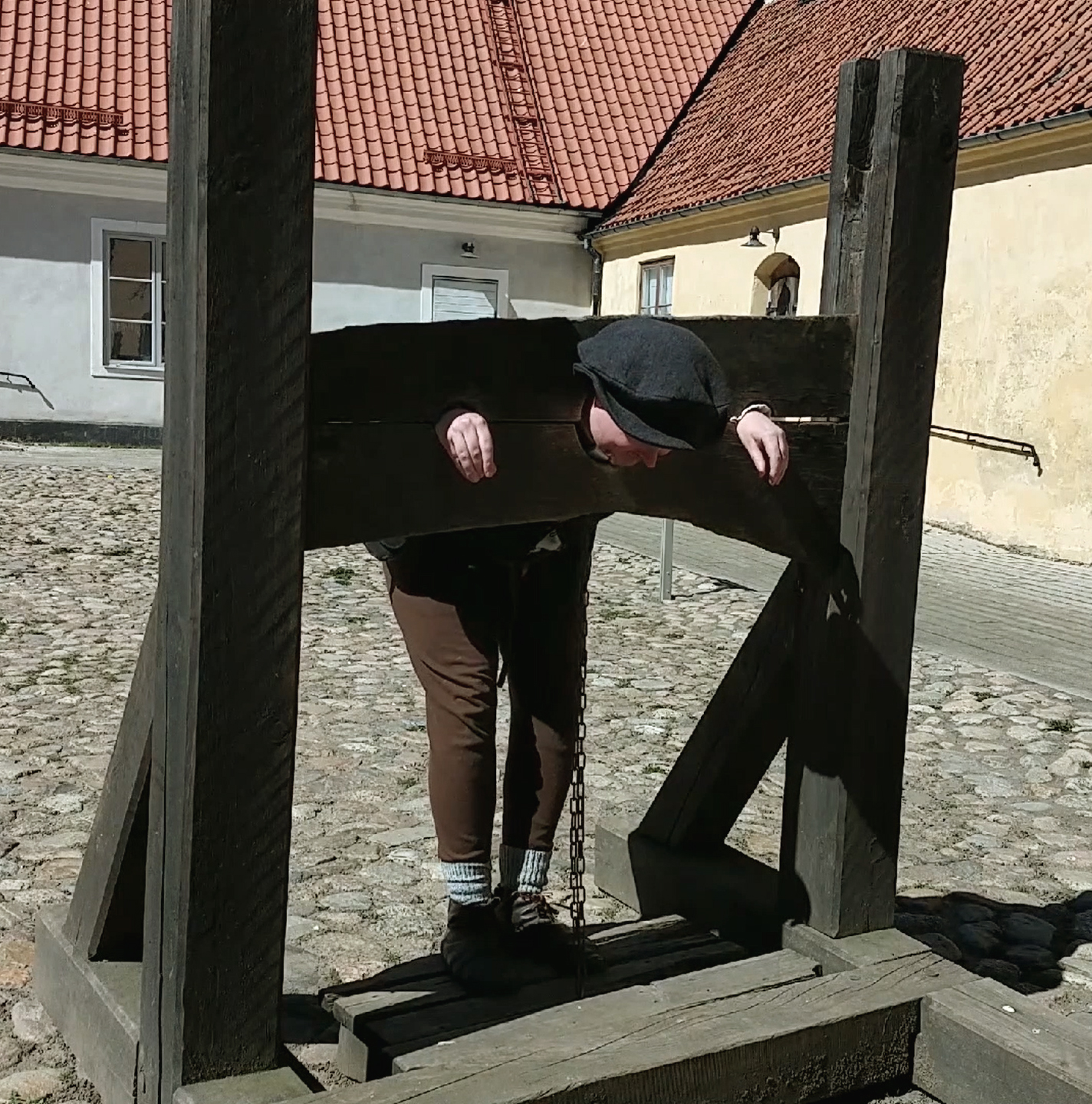 A man in medieval costume locked in the pillory in the castle yard. He is stuck with his hands and head in the log of wood. He has brown pants and a black cap.