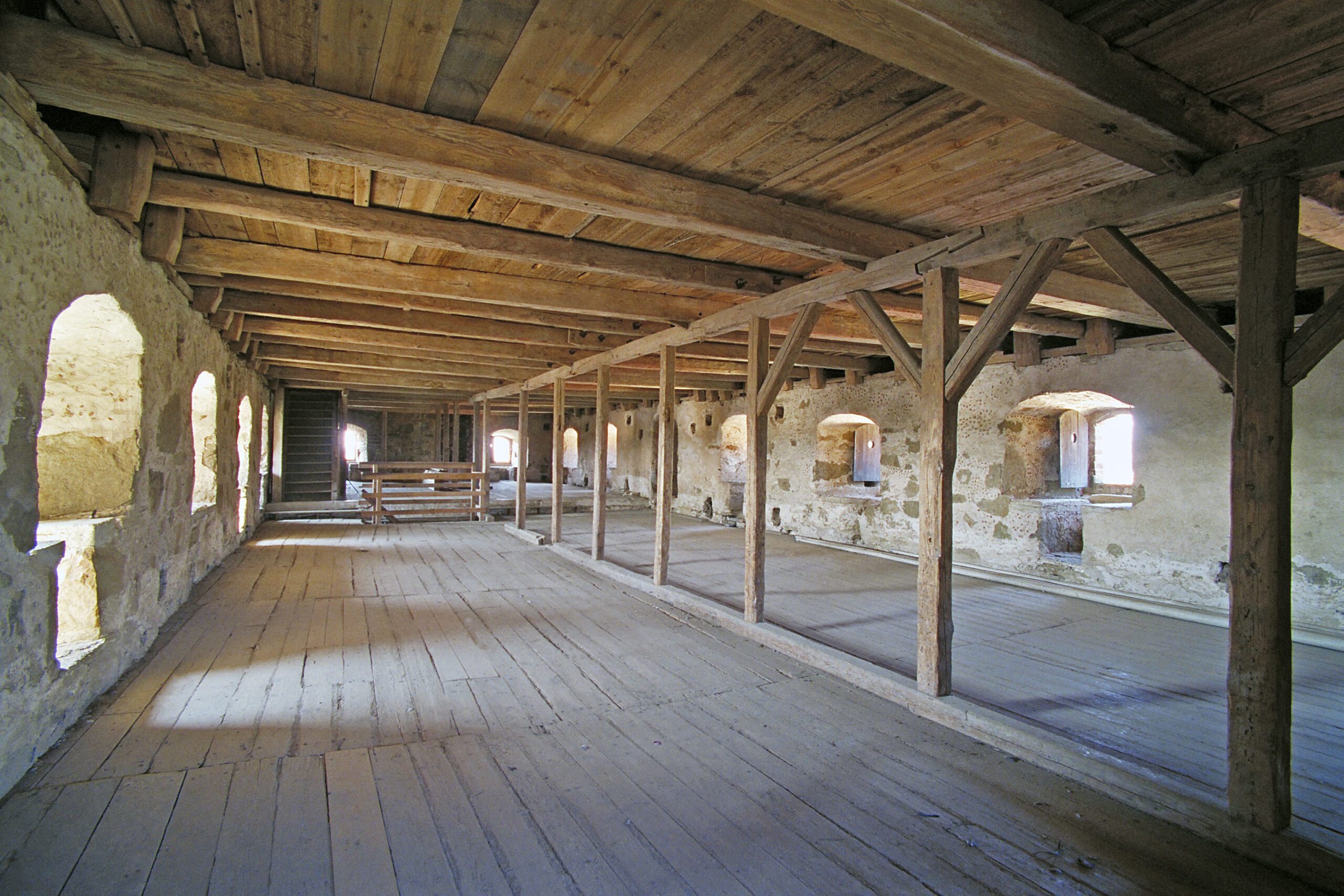 The picture shows an oblong floor plan in the castle with a wooden floor and a wooden ceiling with beams. Along the stone walls are deep window openings.