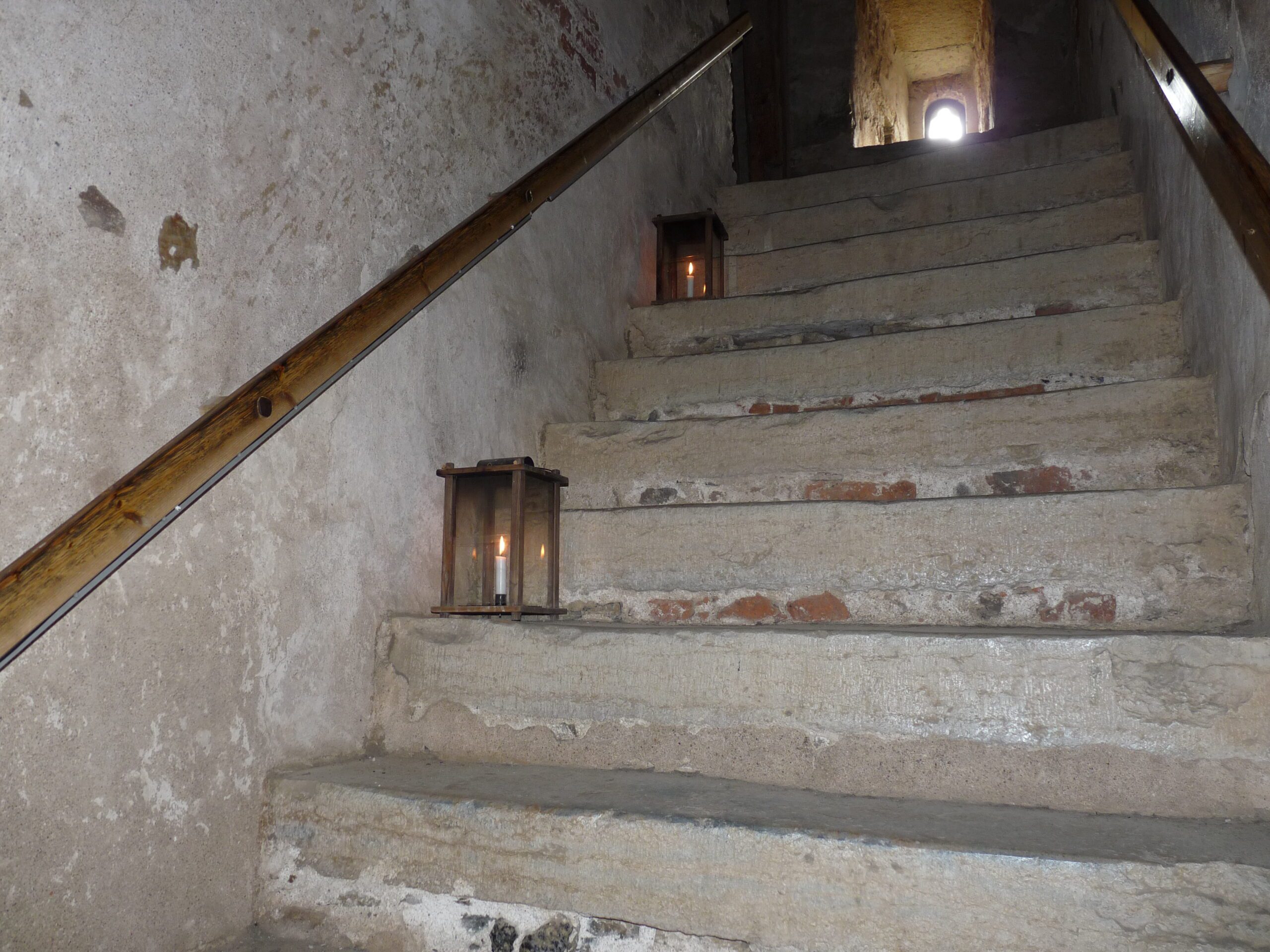 Här ses trappsteg mellan första och andra våningen. På väggen sitter ett räcke och det står ett par lyktor i trappan.
