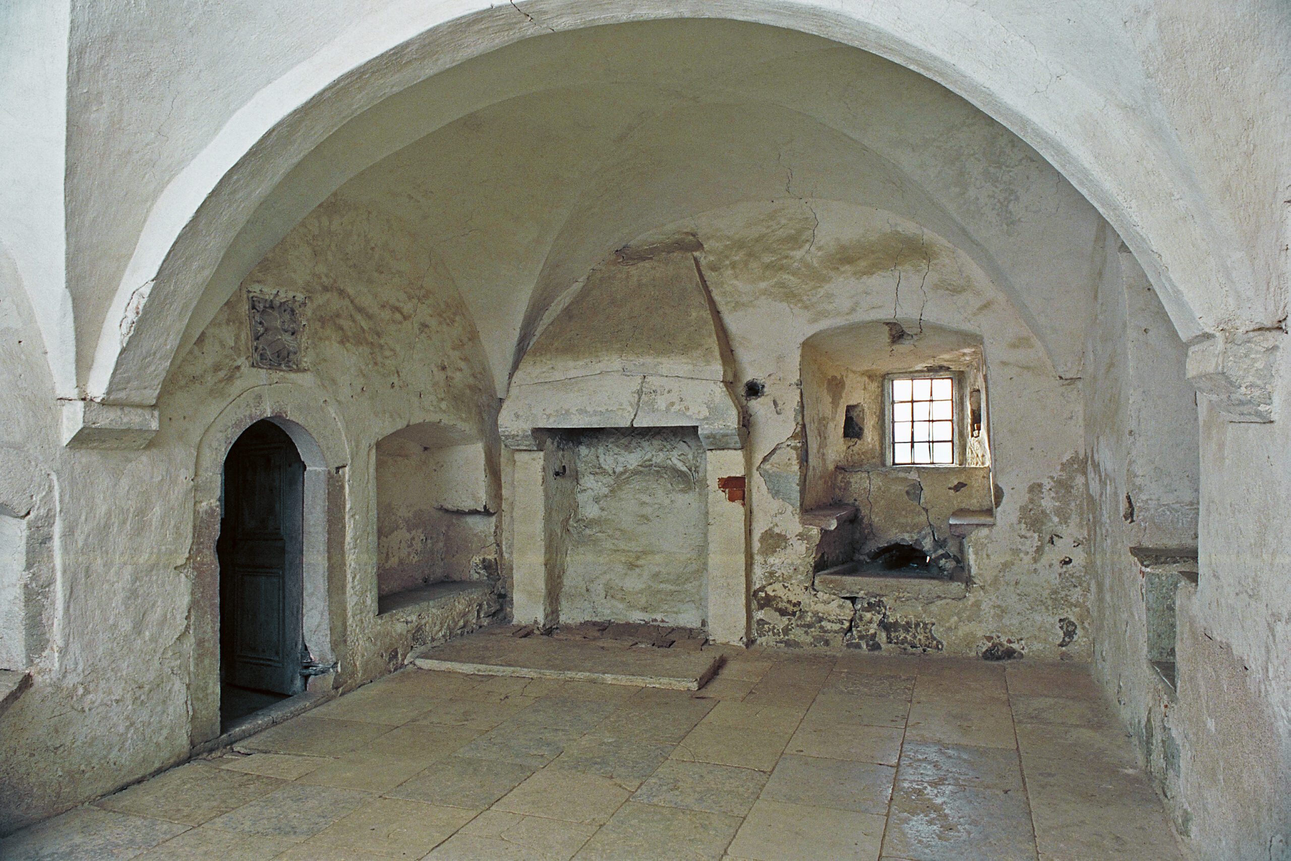 På bilden ses ett medeltida rum i borgen helt byggt i sten. I mitten finns en spis, till vänster en portal och till höger en fönsternisch med sittplatser.
