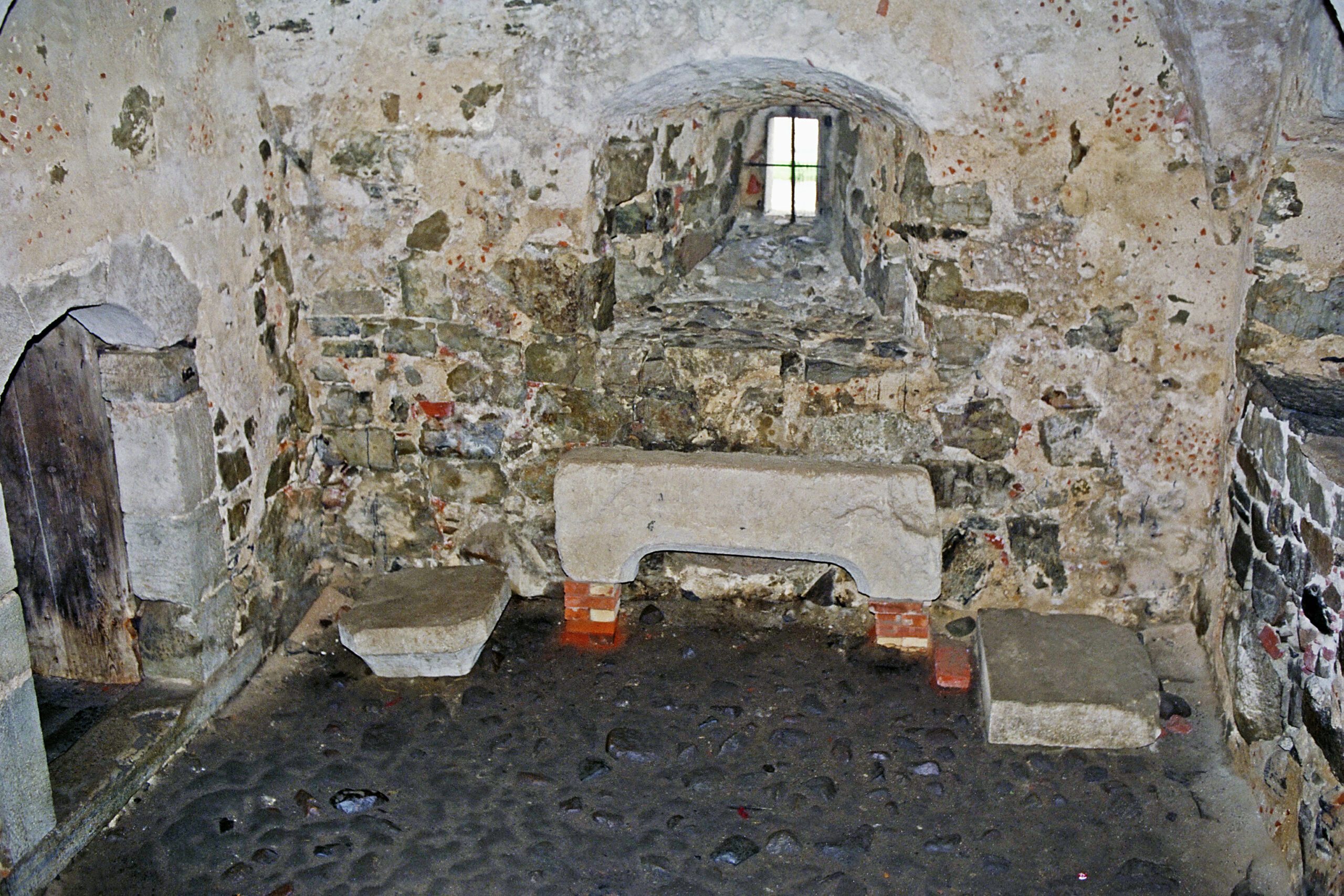 Bilden visar spiskällaren med kullerstensgolv och en fönsternisch. Mot väggen står stenlämningar från borgen.