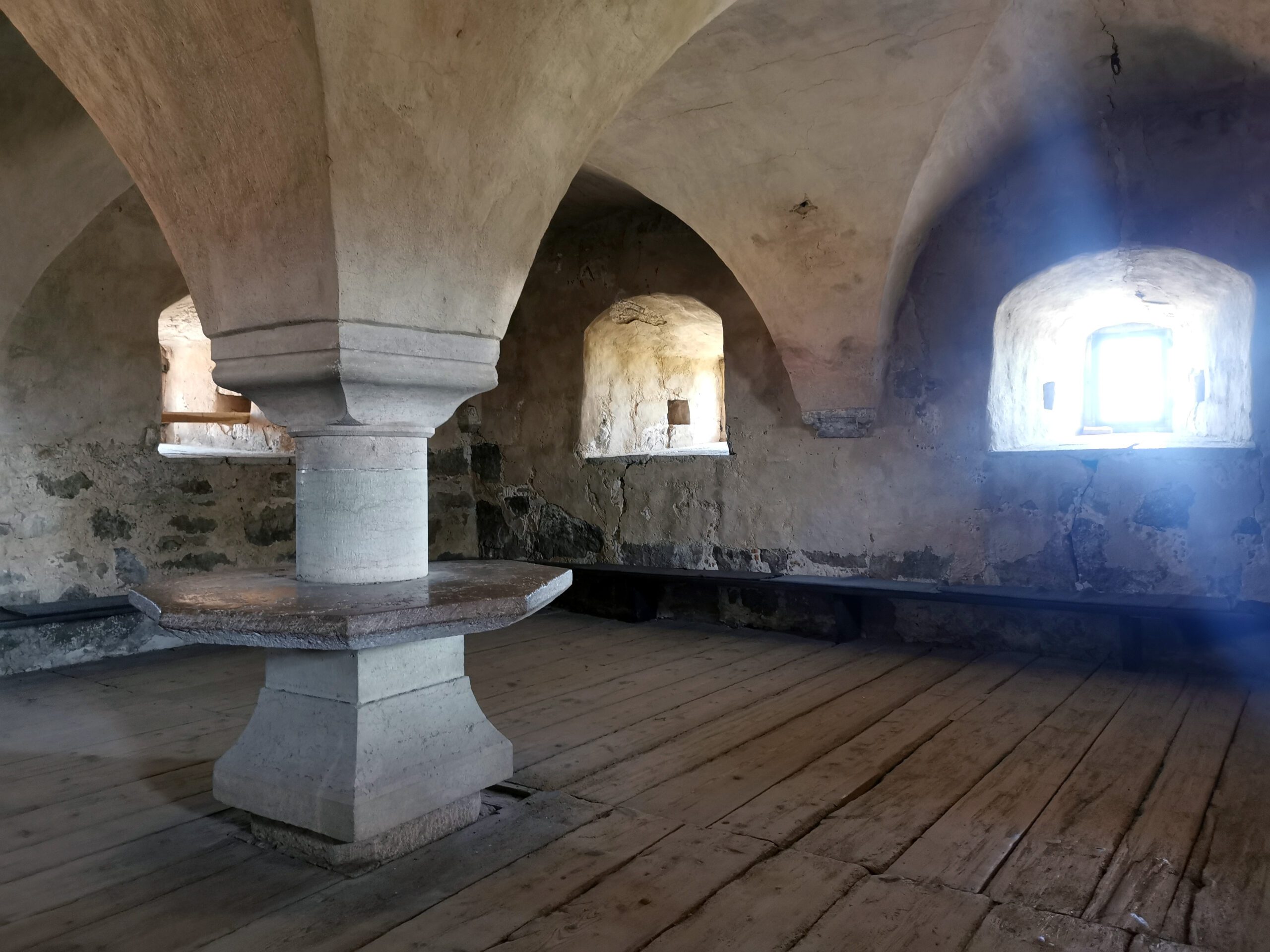 A room with a vaulted ceiling and rough wooden floorboards. In the middle, the vault descends into a pillar with an octagonal table top around it. Fixed benches around the walls and deep window niches.