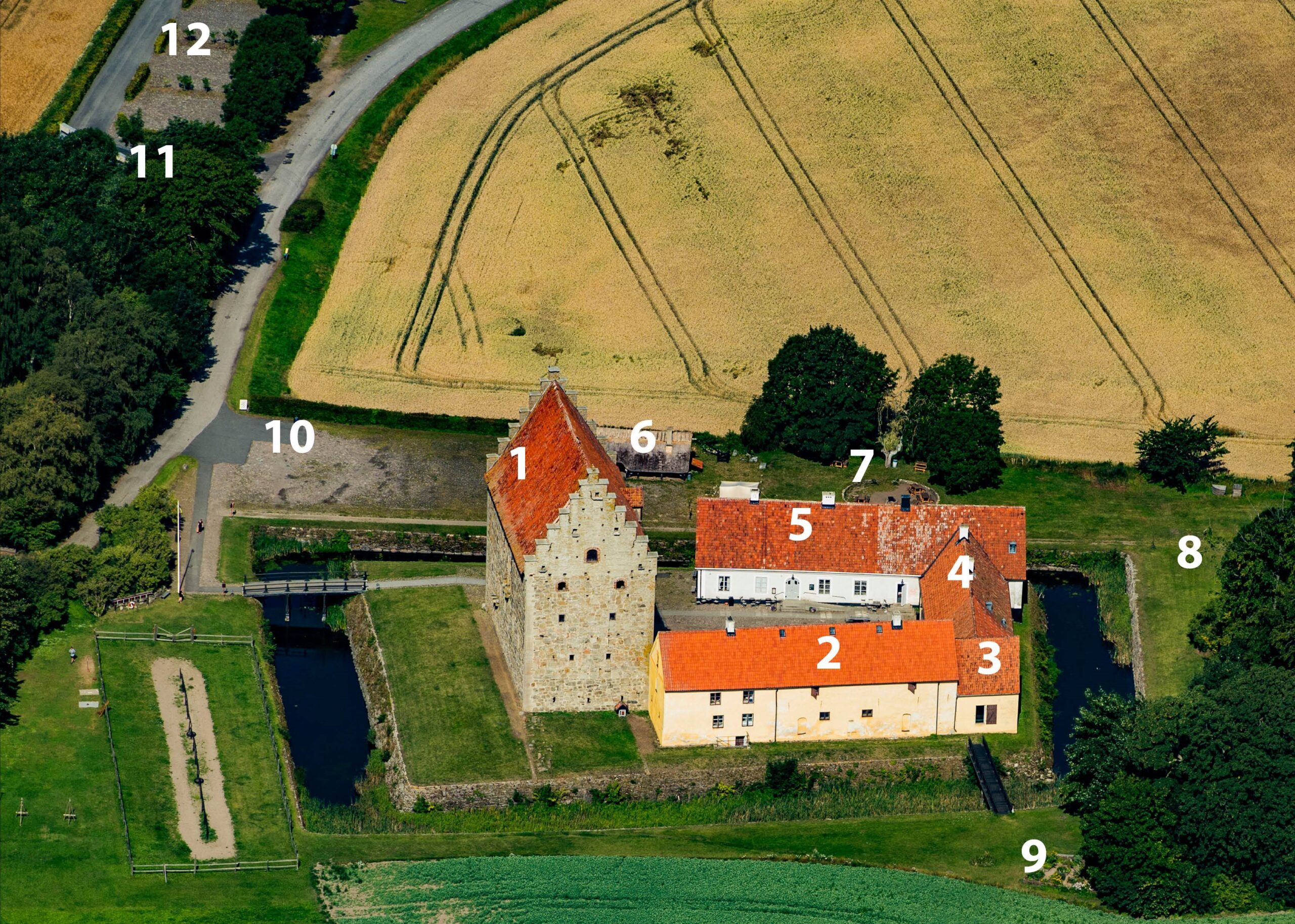 Flygbild över Glimmingehus med borgen och längor i mitten. Bakom dem ses ett åkerfält och framför grönytor.