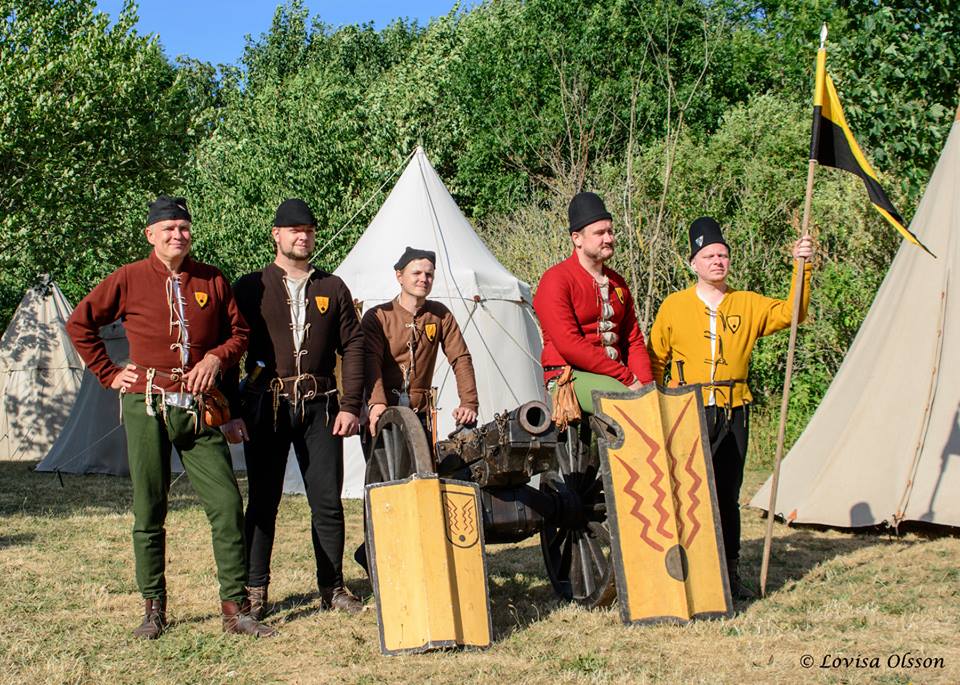 En samling personer i senmedeltida dräkter står utomhus vid en kanon.